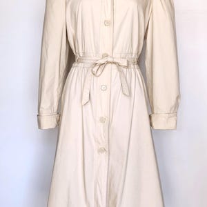 May include: A long-sleeved, cream-colored hooded jacket with a button-up front and a tie belt. The jacket has a slightly flared skirt and is made of a lightweight fabric.