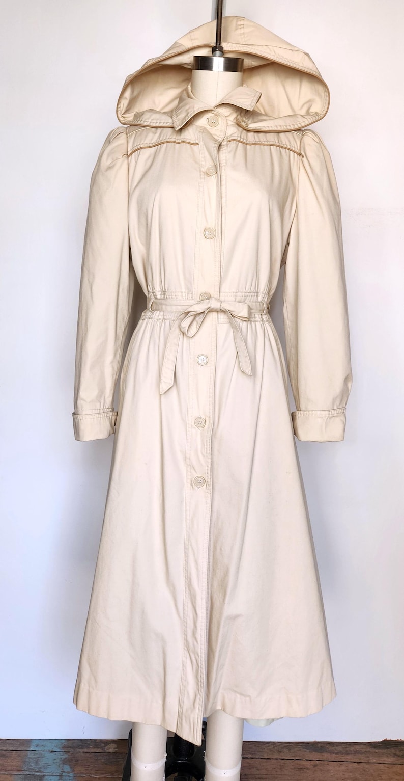 May include: A long-sleeved, cream-colored hooded jacket with a button-up front and a tie belt. The jacket has a slightly flared skirt and is made of a lightweight fabric.