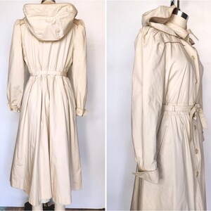May include: A long, white, hooded raincoat with a drawstring waist and a button-up front. The raincoat has a relaxed fit and a slightly flared skirt.