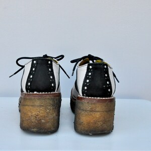 May include: A pair of black and white saddle shoes with a thick platform sole. The shoes have a white leather upper with a black leather panel on the back. The shoes are worn and have a distressed look.
