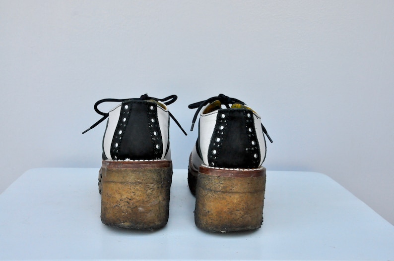 May include: A pair of black and white saddle shoes with a thick platform sole. The shoes have a white leather upper with a black leather panel on the back. The shoes are worn and have a distressed look.