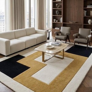 May include: A rectangular area rug featuring a geometric pattern in cream, mustard yellow, and black. The rug has a thick, textured pile and is positioned in a living room with a cream sofa, two gray armchairs, and a stone-topped coffee table.