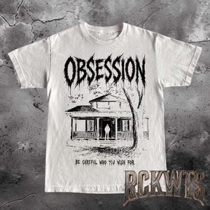 May include: A white short-sleeve t-shirt featuring a black graphic print of a house with a figure standing in the doorway. The word OBSESSION is printed in a jagged font above the house, and the text BE CAREFUL WHO YOU WISH FOR is below.
