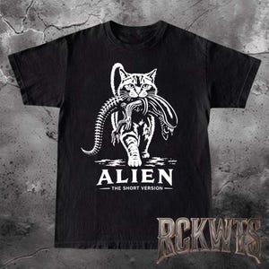 May include: A black short-sleeved t-shirt featuring a white graphic of a cat carrying a xenomorph creature in its mouth. Below the graphic, the text reads ALIEN THE SHORT VERSION. The design is centered on the chest of the garment.