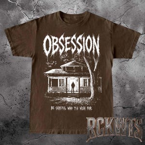 May include: A brown short-sleeved t-shirt featuring a white graphic print of a house with a person standing in the doorway. The word OBSESSION is printed in a jagged font above the house, and the text BE CAREFUL WHO YOU WISH FOR is below.