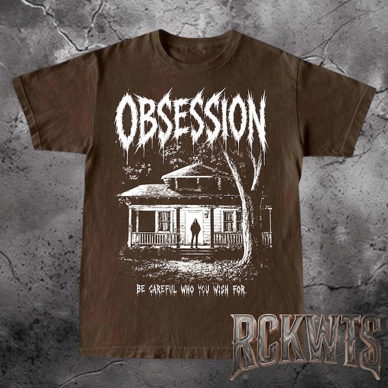May include: A brown short-sleeved t-shirt featuring a white graphic print of a house with a person standing in the doorway. The word OBSESSION is printed in a jagged font above the house, and the text BE CAREFUL WHO YOU WISH FOR is below.