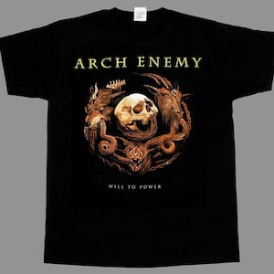 Puede incluir: Una camiseta negra de manga corta con el texto ARCH ENEMY en dorado en la parte superior y WILL TO POWER en la inferior. El gráfico central muestra un cráneo humano rodeado por una cabeza de lobo, una de cabra y una de jabalí en tonos marrones.