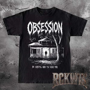 May include: A black short-sleeved t-shirt featuring a white graphic of a house with a figure standing in the doorway. The text "OBSESSION" appears at the top in a jagged font, and the text "BE CAREFUL WHO YOU WISH FOR." is printed below the house.