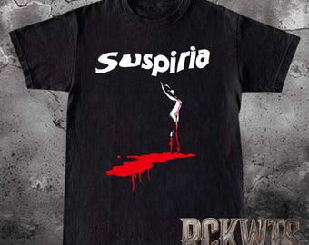Puede incluir: Una camiseta negra de manga corta con el texto blanco Suspiria impreso en el pecho. Debajo del texto hay un gráfico de la silueta blanca de una persona de pie sobre un charco rojo de líquido que se asemeja a una salpicadura en la tela.
