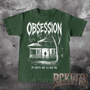 May include: A dark forest green short-sleeve t-shirt featuring a white graphic print of a house with a figure standing in the doorway. The word OBSESSION is printed in a jagged font above, and the text BE CAREFUL WHO YOU WISH FOR is below.