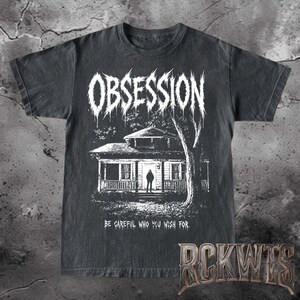 May include: A black short-sleeved t-shirt featuring a white graphic print of a house with a figure standing in the doorway. The word OBSESSION is printed in a jagged font above the house, and the text BE CAREFUL WHO YOU WISH FOR is below.