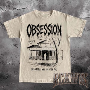 May include: A beige short-sleeve t-shirt featuring a black graphic print of a house with a figure standing in the doorway. The word OBSESSION is printed in a jagged font above the house, and the text BE CAREFUL WHO YOU WISH FOR is below.