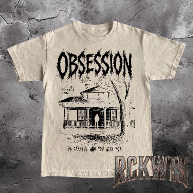 May include: A beige short-sleeve t-shirt featuring a black graphic print of a house with a figure standing in the doorway. The word OBSESSION is printed in a jagged font above the house, and the text BE CAREFUL WHO YOU WISH FOR is below.