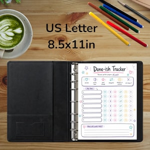May include: A black binder containing a Done-ish Tracker page with US Letter 8.5x11in dimensions. The page features task lists and status icons. Nearby are a cup of green matcha, five colored pens, and a pencil on a wooden surface.