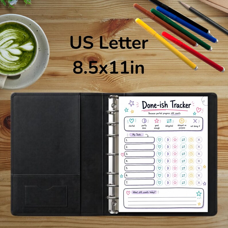 May include: A black binder containing a Done-ish Tracker page with US Letter 8.5x11in dimensions. The page features task lists and status icons. Nearby are a cup of green matcha, five colored pens, and a pencil on a wooden surface.