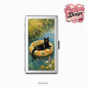 May include: A rectangular silver metal case featuring a printed illustration of a black cat resting on an orange-patterned inflatable pool float in a pond surrounded by colorful wildflowers and green foliage, with a pink heart logo in the corner.