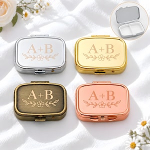 Pode incluir: Quatro porta-comprimidos retangulares de metal com acabamentos em prata, ouro, bronze antigo e ouro rosa. Cada estojo apresenta uma gravação das letras A+B sobre um ramo floral. Um detalhe mostra um estojo aberto com espelho e dois compartimentos.
