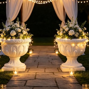 May include: Two large, white, ornate stone urns filled with white flowers and greenery, wrapped in warm glowing string lights. The urns sit on a stone pathway, flanked by small glass candle holders with lit candles, leading toward a draped archway.