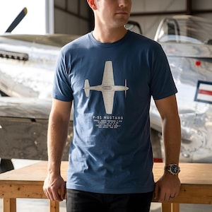 May include: A blue short-sleeve t-shirt featuring a white line drawing of a P-51 Mustang airplane viewed from above. Below the graphic, text lists the model, wingspan of 37 ft 0 in, length of 32 ft 3 in, engine, and max speed of 437 mph.