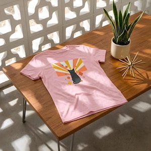 May include: A light pink short-sleeve t-shirt featuring a graphic print of a black cat centred against a background of orange, yellow, and white sunburst rays, accented with small colourful diamond shapes and starburst icons.