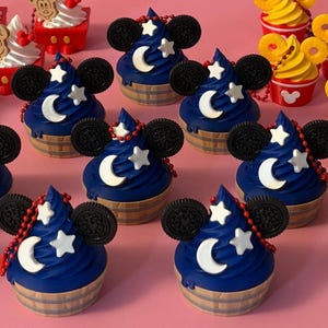 May include: A collection of miniature cupcake-shaped charms featuring dark blue frosting, two black circular cookie ears, a white crescent moon, and a white star. Each charm sits in a tan base and includes a red beaded chain attachment.