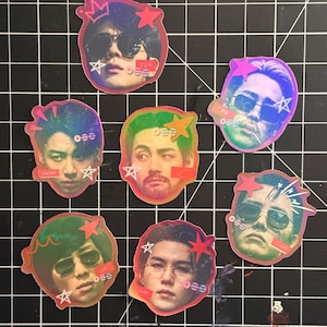 May include: Seven holographic stickers featuring portraits of individuals wearing sunglasses, accented with red and pink star graphics, small circular icons, and handwritten-style text labels including names like Jin, Jimin, and Suga.