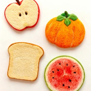 Op de afbeelding: Vier naaldgevilte etenswaren: een rood met crèmekleurig appelschijfje met bruine pitjes, een oranje pompoen met groene blaadjes, een snee goudbruin geroosterd brood en een ronde watermeloen met rood vruchtvlees, zwarte pitjes en een groene schil.