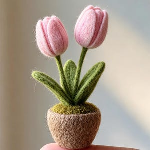 Könnte beinhalten: Ein handgefertigtes Objekt aus Nadelfilz, bestehend aus zwei zartrosa Tulpenblüten mit grünen Stielen und Blättern in einem kleinen, strukturierten braunen Topf mit moosartiger Oberfläche. Das gesamte Stück ist miniaturisiert und hat eine flauschige Textur.
