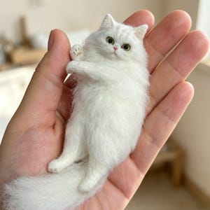 Puede incluir: Una figura miniatura de gato blanco hecha con fieltro de aguja, con pelaje largo y esponjoso, orejas puntiagudas y ojos claros. El pequeño gato artesanal está representado en posición reclinada con una pata levantada, mostrando textura.