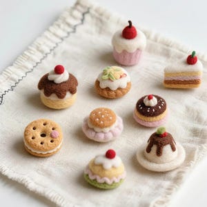 May include: A collection of nine miniature needle-felted desserts including cupcakes, cookies, and cake slices. Each piece features soft textures in shades of brown, pink, white, and green, with small red berry accents on several of the items.
