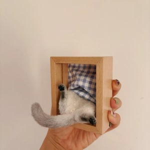 May include: A needle-felted grey and white cat figurine positioned inside a small, rectangular light-coloured wooden frame. The cat is shown from the rear, with its tail extended and its paws visible, resting against a blue and white gingham fabric.
