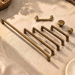 May include: A collection of antique brass-toned metal cabinet hardware, including five long bar pulls and one smaller handle, each engraved with names like Noah, Lily, Owen, Mia, and Leo, alongside two round knobs engraved with the name Zoe.