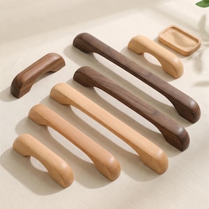 Peut inclure: Une collection de six poignées de placard en bois de longueurs variées et deux tons de bois distincts, incluant du bois clair naturel et du bois brun foncé, disposées à côté d'un petit plateau rectangulaire en bois sur une surface neutre.