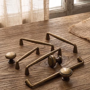Op de afbeelding: Een collectie antiek messing beslag, bestaande uit drie langwerpige ladehandgrepen met gebogen stangen en twee ronde kastknoppen. Elk stuk heeft een verweerde, metalen afwerking met donkere accenten en een glad, afgerond oppervlak.