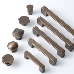 May include: A collection of antique brass cabinet hardware including four rectangular drawer pulls and five knobs in various shapes, including round, hexagonal, and square designs, all featuring a brushed metallic finish and smooth edges.