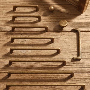 May include: A collection of antique brass cabinet hardware including seven horizontal bar pulls of varying lengths, one vertical bar pull, and two round knobs. The metal hardware features a weathered, textured finish with a warm golden-brown tone.