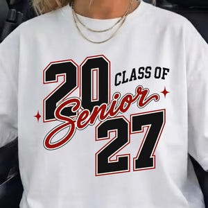 May include: A white short-sleeved t-shirt featuring a graphic print that reads 20 CLASS OF 27. The numbers 20 and 27 are in large black block font with red outlines, while the word Senior is in a red cursive script with black outlines.