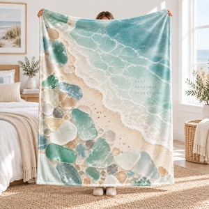 May include: A soft throw blanket featuring a watercolor-style print of a sandy beach with smooth, rounded stones in shades of teal, green, and beige along the shoreline. The text "SALT RESTORES EVERYTHING" is printed in white on the water.