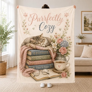 Puede incluir: Una manta de forro polar color crema con el texto "Purrfectly Cozy" sobre una ilustración de un gato atigrado durmiendo sobre tres libros titulados "Feline Tales", "A Cat's Life" y "Paws & Pages", junto a un jarrón con flores y unas gafas.