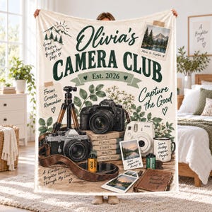May include: A cream-colored throw blanket featuring the text Olivia's Camera Club Est. 2026, Capture the Good, and Focus Create Remember. The design includes illustrations of a black DSLR camera, a vintage camera, a tripod, a white instant camera, a mug, film rolls, and stacked books with nature-themed accents.