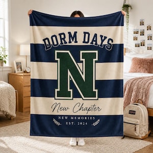 以下が含まれることがあります： ネイビーブルーとクリーム色のストライプ柄フリースブランケット。カレッジフォントで「DORM DAYS」の文字、中央に大きなダークグリーンの「N」、スクリプト体とセリフ体で「New Chapter NEW MEMORIES EST. 2024」の文字がプリントされています。