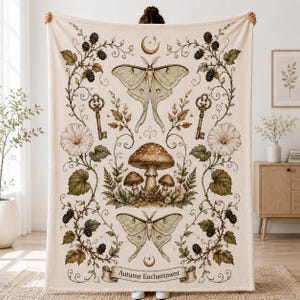 Könnte beinhalten: Eine cremefarbene Wohndecke mit botanischem und himmlischem Aufdruck. Das Design zeigt zwei große Nachtfalter, drei Pilze, zwei antike Schlüssel, Brombeerranken und Blumen. Am unteren Rand steht der Text "Autumn Enchantment".