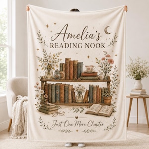 May include: A cream-colored fleece blanket featuring a printed illustration of two wooden bookshelves filled with vintage books, floral arrangements, and a teacup. The text reads Amelia's READING NOOK and Just One More Chapter with stars and a moon.