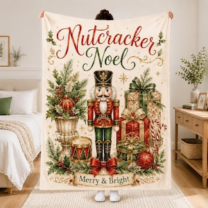 May include: A cream-colored throw blanket featuring a central nutcracker illustration, surrounded by Christmas gifts, a drum, and greenery. The text reads Nutcracker Noel at the top and Merry & Bright on a banner at the bottom, with red and green accents.