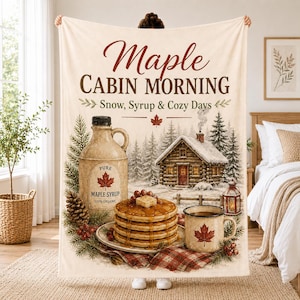 Pode incluir: Uma manta de lã cor de creme com o texto Maple CABIN MORNING Snow, Syrup & Cozy Days. O design inclui uma garrafa de xarope de ácer, uma pilha de panquecas com xarope, uma cabana de madeira numa floresta nevada e uma caneca com uma folha.