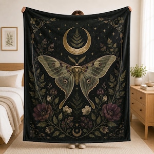 May include: A black rectangular throw blanket featuring a large central moth with muted green and brown wings. The design includes a golden crescent moon containing a fern leaf, surrounded by dark purple flowers, green foliage, and small stars.