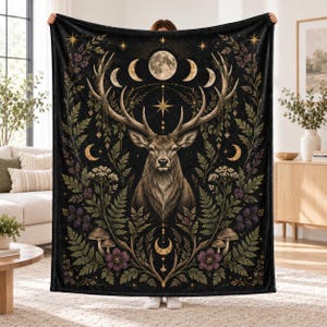 May include: A black rectangular throw blanket featuring a central illustration of a deer head with large antlers. The design includes a moon phase cycle, stars, ferns, purple flowers, mushrooms, and berries arranged in a symmetrical pattern.