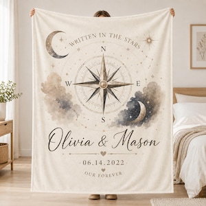 May include: A cream-colored fleece blanket featuring a central compass rose design, celestial moon and star motifs, and watercolor-style clouds. The text reads "WRITTEN IN THE STARS", "Olivia & Mason", "06.14.2022", and "OUR FOREVER" in black script and print.