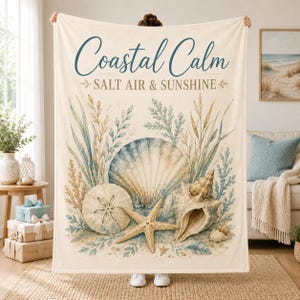May include: A cream-coloured throw blanket featuring a watercolour illustration of seashells, a starfish, and sea grass. The text "Coastal Calm" appears in blue script above the words "SALT AIR & SUNSHINE" in a dark blue serif font.