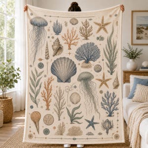 May include: A cream-coloured throw blanket featuring a detailed marine life pattern. The design includes illustrations of blue jellyfish, various seashells, orange and blue coral, starfish, and seaweed arranged across the fabric surface.